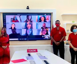 Tres ejecutivos de UAGM parados frente a un televisor donde se proyectan un grupo de estudiantes virtual