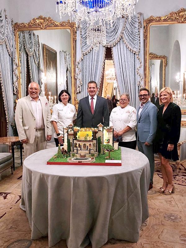 grupo de asociados junto la gobernador exhibiendo Castillo Serrallés en bizcocho